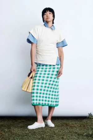 Model standing against a white backdrop wearing a white top over a blue gingham shirt, green gingham skirt, and holding a yellow handbag