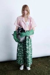 Model in pink shirt and green patterned pants wearing long suede gloves holding a patterned bag against a studio backdrop.