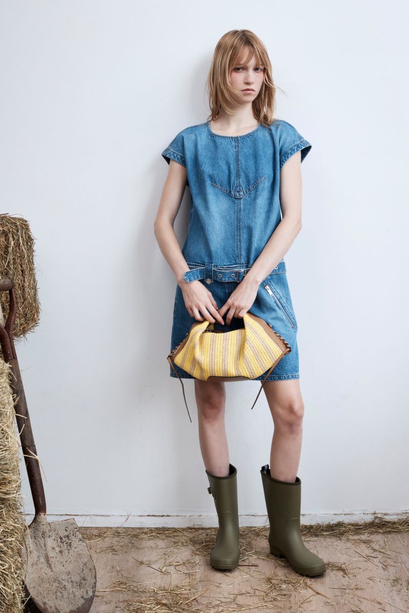Model wearing a blue denim dress and green boots holding a yellow handbag against a white wall with straw on the floor.