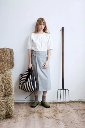 Model wearing a white top and grey skirt standing next to hay bales and a pitchfork against a white wall.