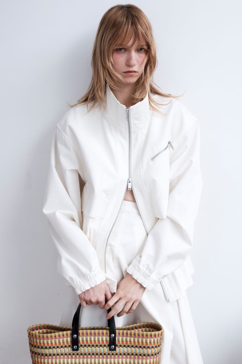 Model wearing a white zip-up jacket holding a woven basket bag against a plain white background.