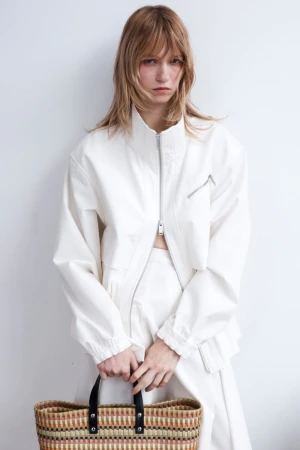 Model wearing a white zip-up jacket holding a woven basket bag against a plain white background.