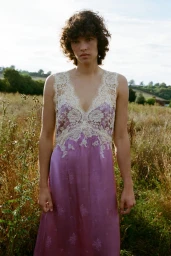 Model with dark curly hair wearing a purple slip dress with cream lace trim standing in an outdoor meadow.