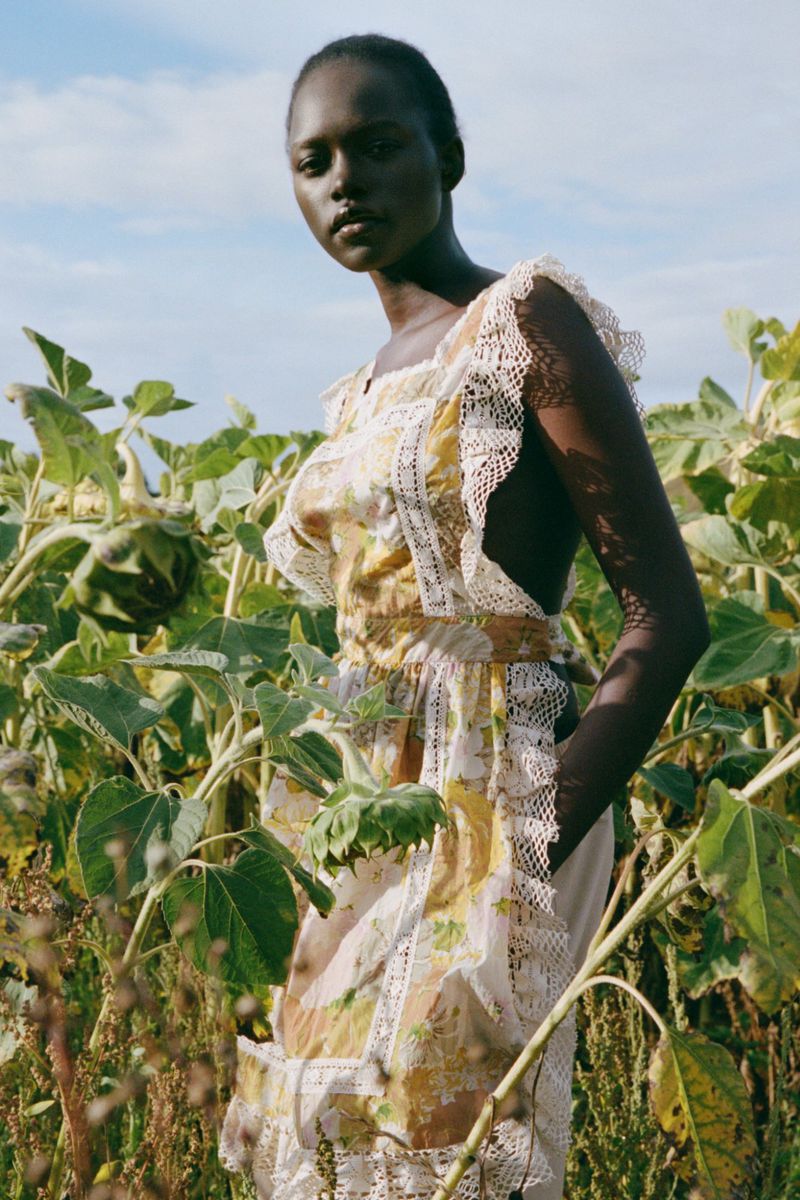 Model wearing a ruffled floral dress standing among tall green plants under an open sky