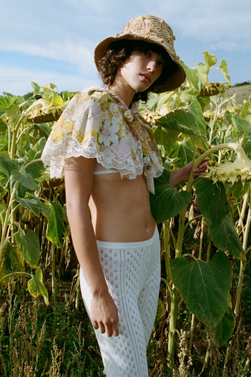 Model standing among tall sunflowers wearing a floral lace capelet, straw bucket hat, and white knit trousers.
