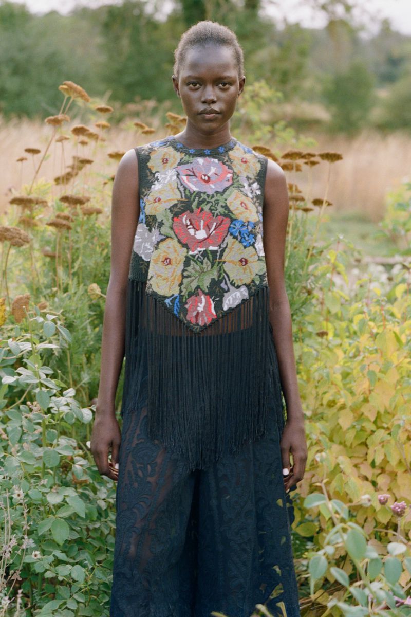 Model wearing a black sleeveless top with a floral embroidered panel and fringe standing in a garden.