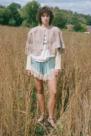 Model standing in a dry grassy meadow wearing a tiered fringed cape and pale blue shorts with lace trim.