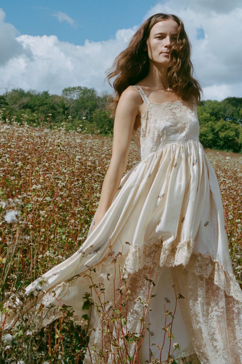 Model in a cream lace-trimmed dress standing in a wildflower field under a partly cloudy sky.