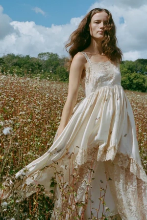 Model in a cream lace-trimmed dress standing in a wildflower field under a partly cloudy sky.