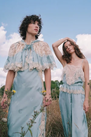 Two female models in light blue satin and lace outfits posing outdoors in a grassy field under a cloudy sky.