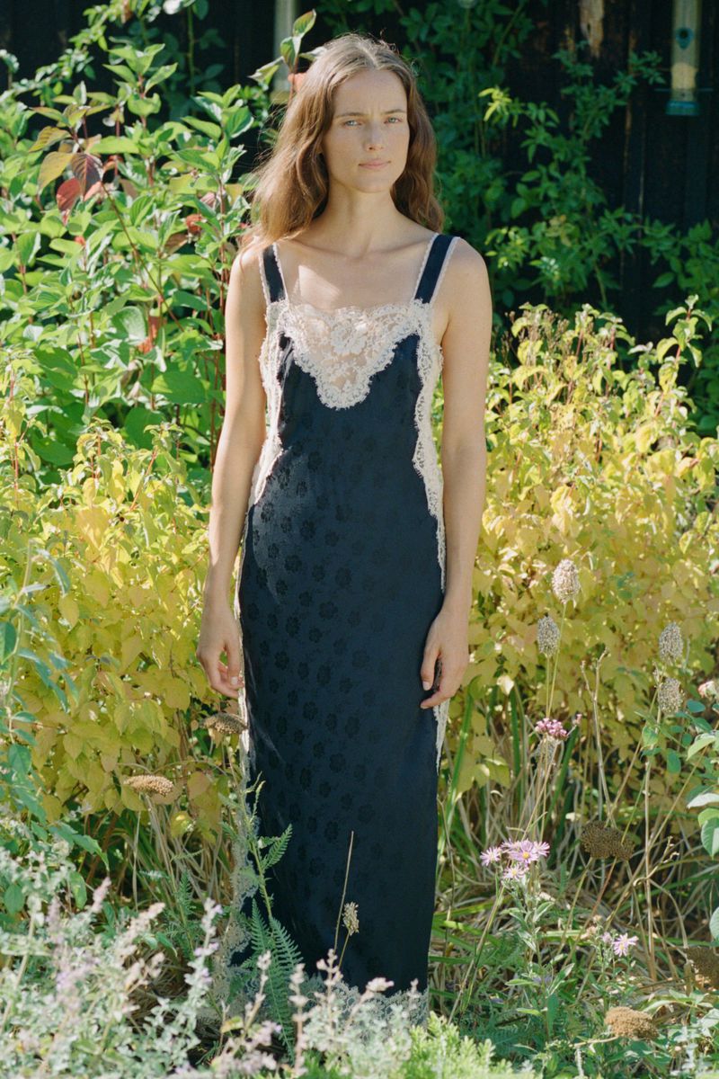 Model standing outdoors in a dark slip dress with lace trim, surrounded by green foliage and garden plants.