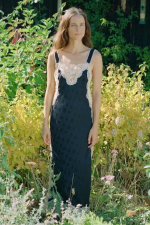 Model standing outdoors in a dark slip dress with lace trim, surrounded by green foliage and garden plants.