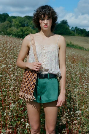 Model with curly dark hair standing in a meadow wearing a floral camisole, green shorts, and carrying a woven patterned bag