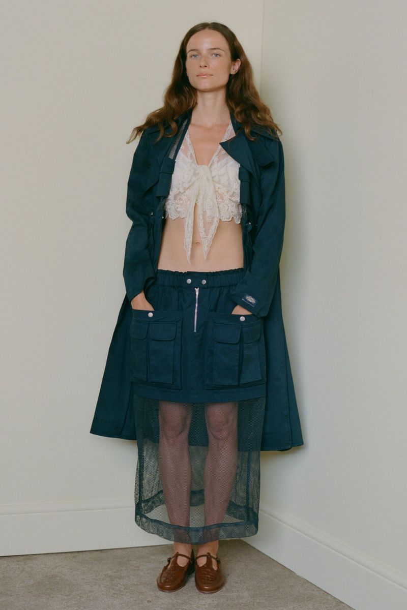 Model standing against a plain corner wall wearing a dark teal coat, lace crop top, utility skirt, and brown shoes.