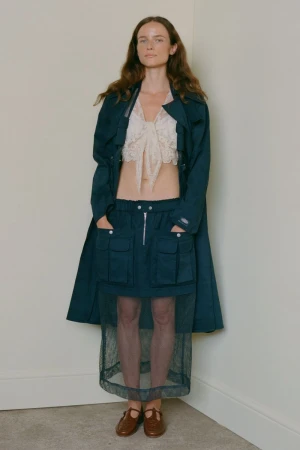 Model standing against a plain corner wall wearing a dark teal coat, lace crop top, utility skirt, and brown shoes.