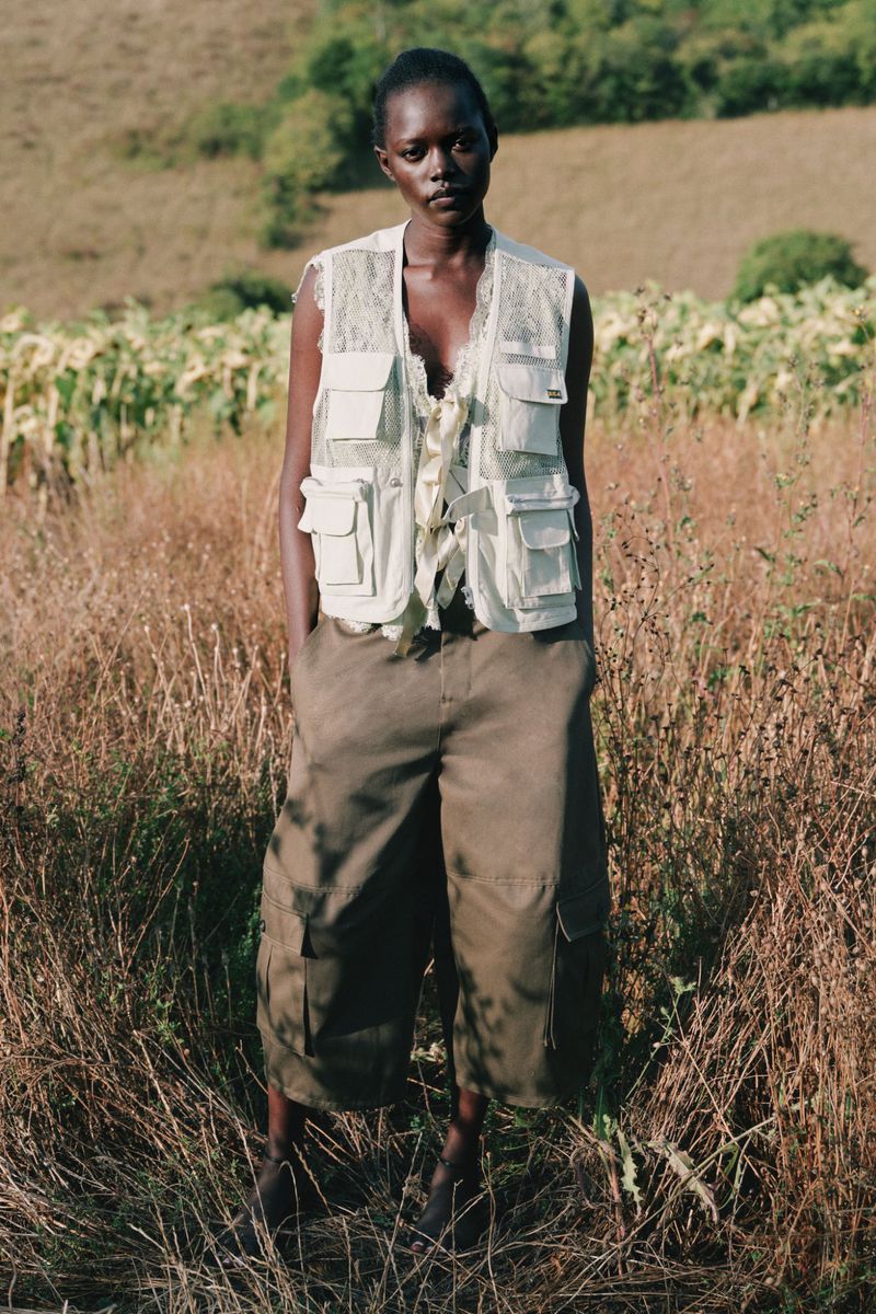 Model wearing a utility vest and cargo shorts standing outdoors in a grassy meadow for the Sea SS27 lookbook.