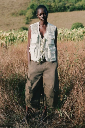 Model wearing a utility vest and cargo shorts standing outdoors in a grassy meadow for the Sea SS27 lookbook.