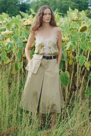 Model standing outdoors in a field of tall sunflowers wearing a patterned camisole and khaki midi skirt for Sea SS27 Lookbook.