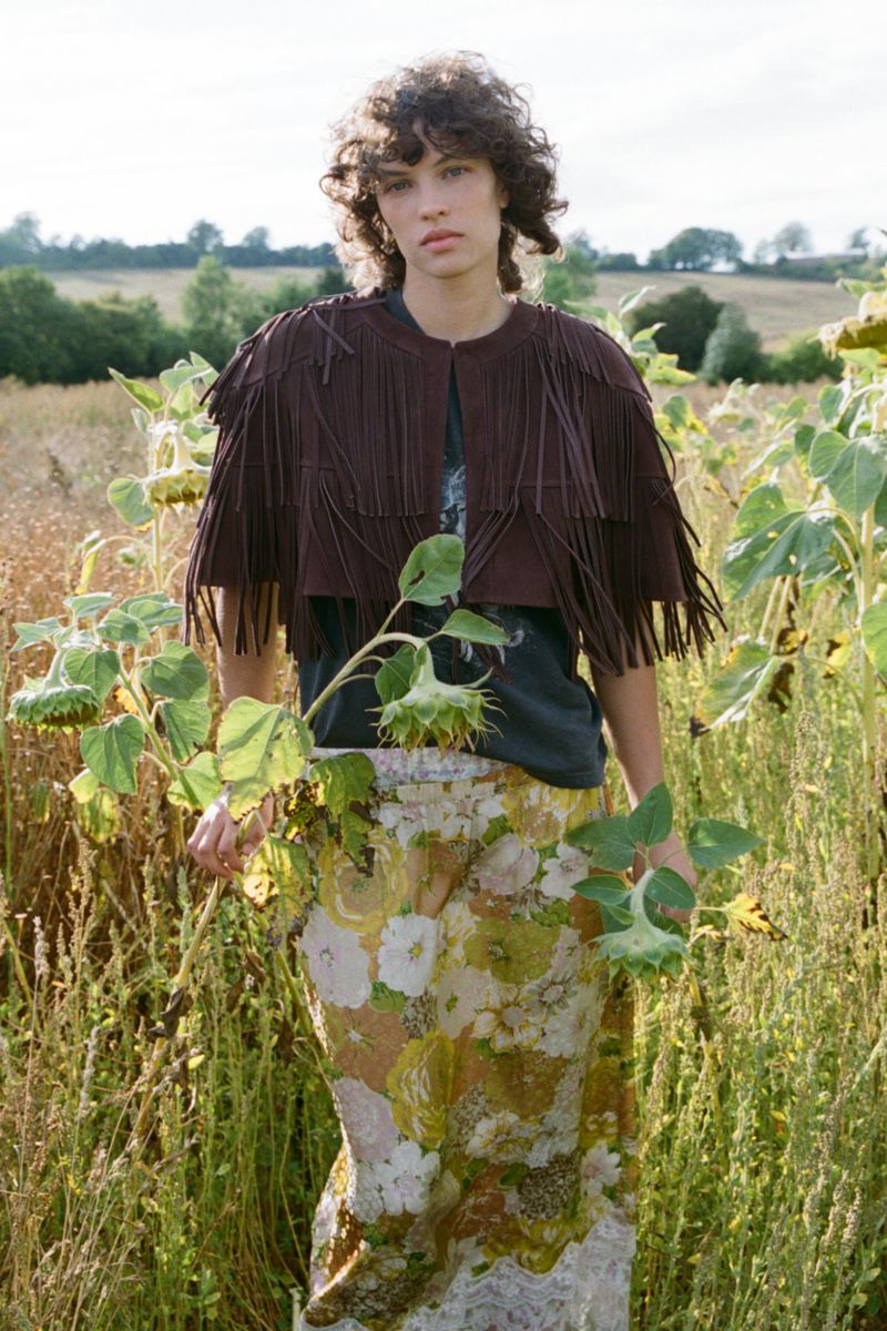 Model standing in a field of sunflowers wearing a fringed brown cape and patterned skirt for Sea SS27 Lookbook.