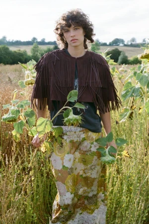 Model standing in a field of sunflowers wearing a fringed brown cape and patterned skirt for Sea SS27 Lookbook.