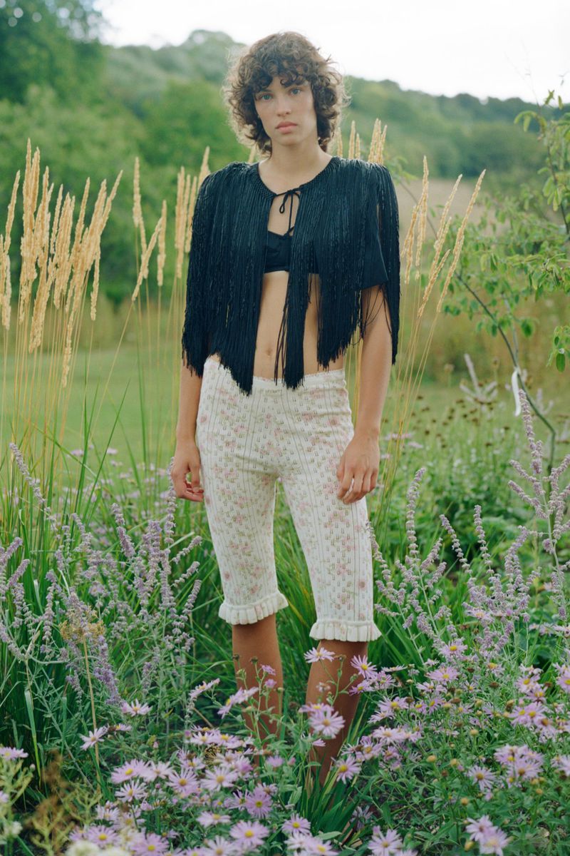 Model standing outdoors in a lush meadow wearing a black fringe top and patterned knit shorts.