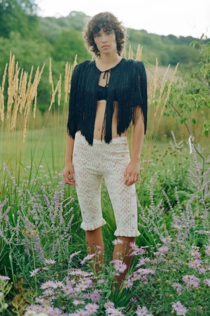 Model standing outdoors in a lush meadow wearing a black fringe top and patterned knit shorts.