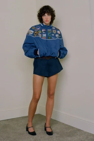 Model in blue patch-embroidered windbreaker and dark shorts posing against a neutral studio wall.