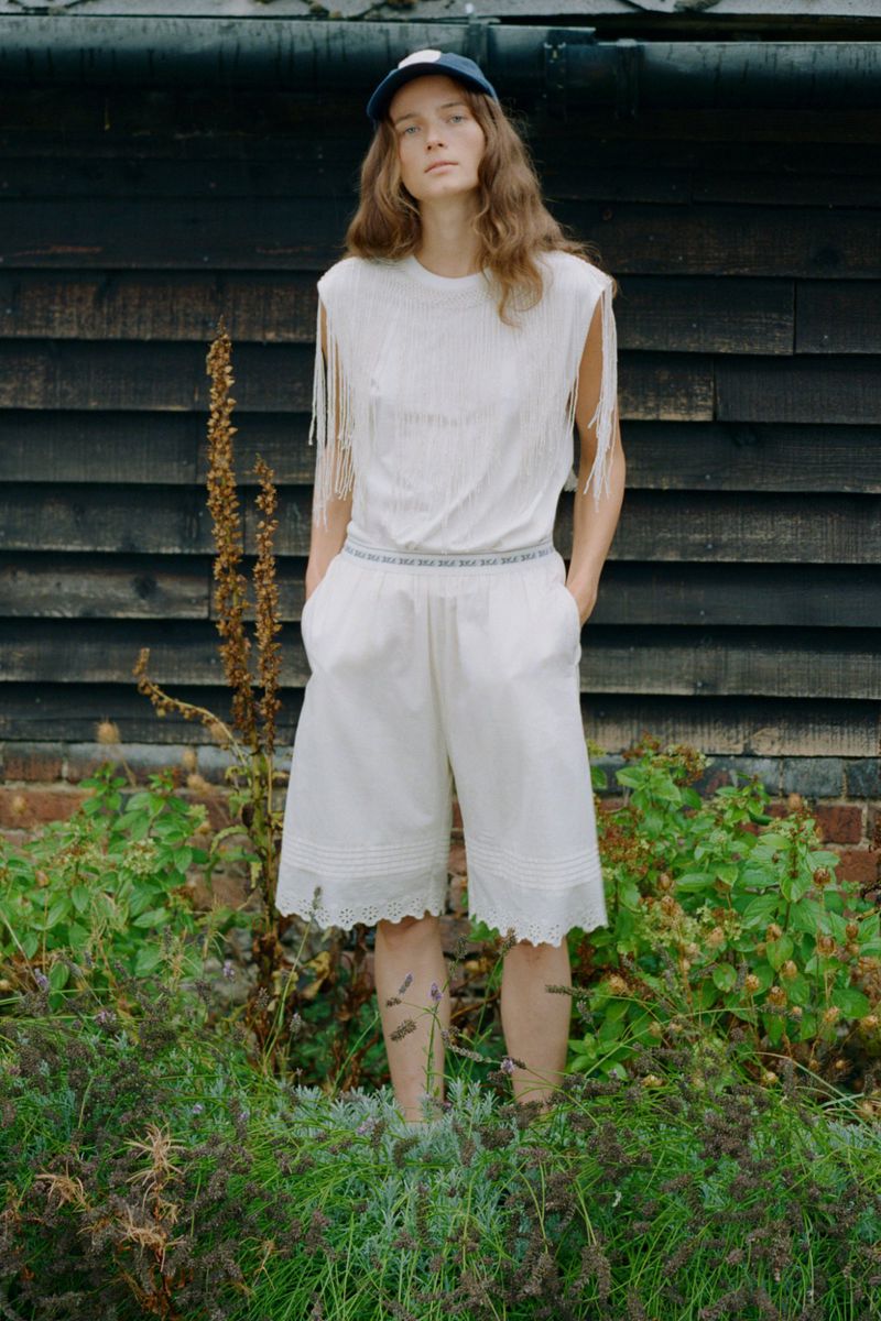 Model standing outdoors wearing a white fringed top and white shorts against dark timber wall