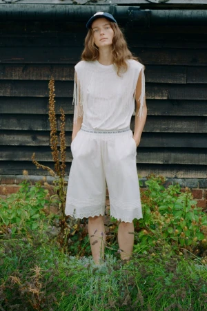 Model standing outdoors wearing a white fringed top and white shorts against dark timber wall