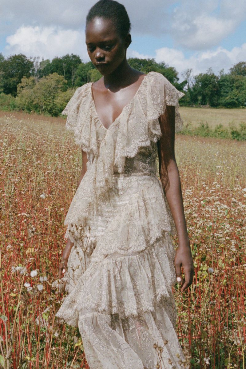 Black model in a cream lace ruffle dress standing in a wildflower field under a partly cloudy sky for Sea SS27 lookbook.