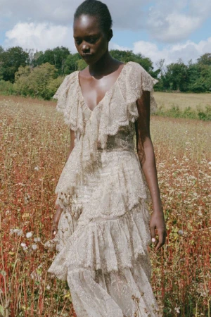 Black model in a cream lace ruffle dress standing in a wildflower field under a partly cloudy sky for Sea SS27 lookbook.