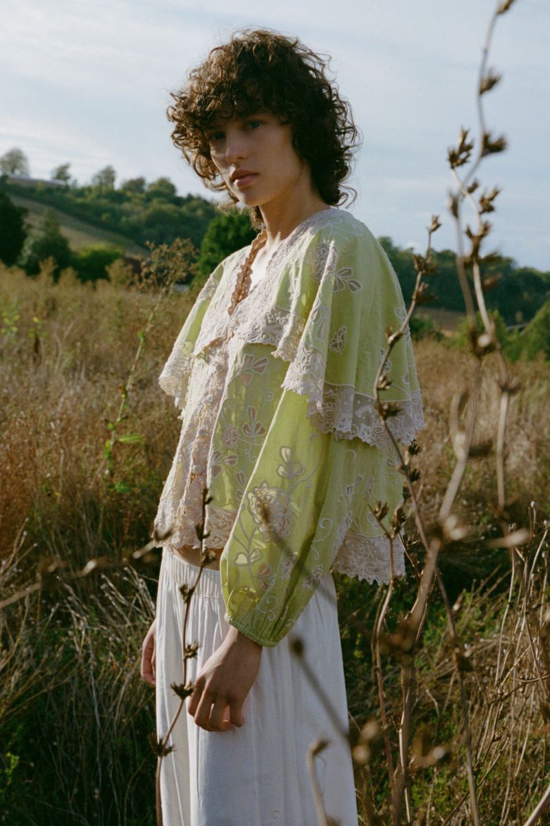Model with curly hair wearing a pale green embroidered top in an outdoor grassy field for Sea SS27 lookbook