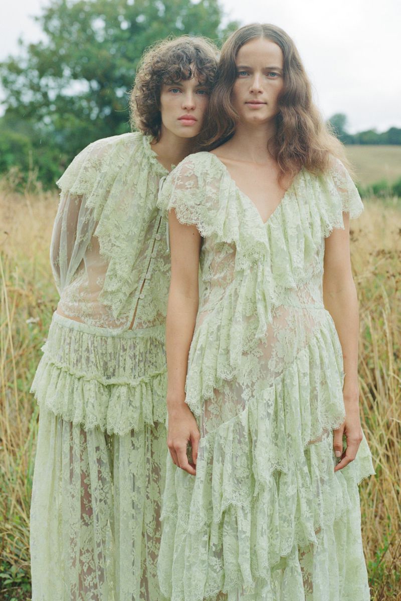 Two female models wearing pale green lace dresses standing in an outdoor meadow field.