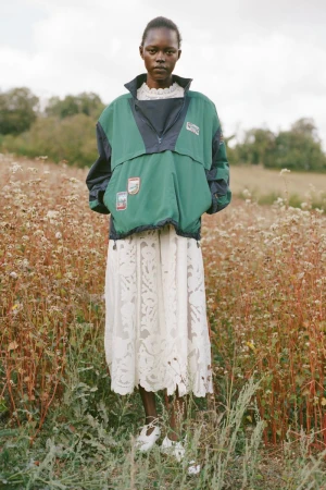 Model standing in a meadow wearing a green pullover windbreaker and white patterned skirt for Sea SS27 lookbook.