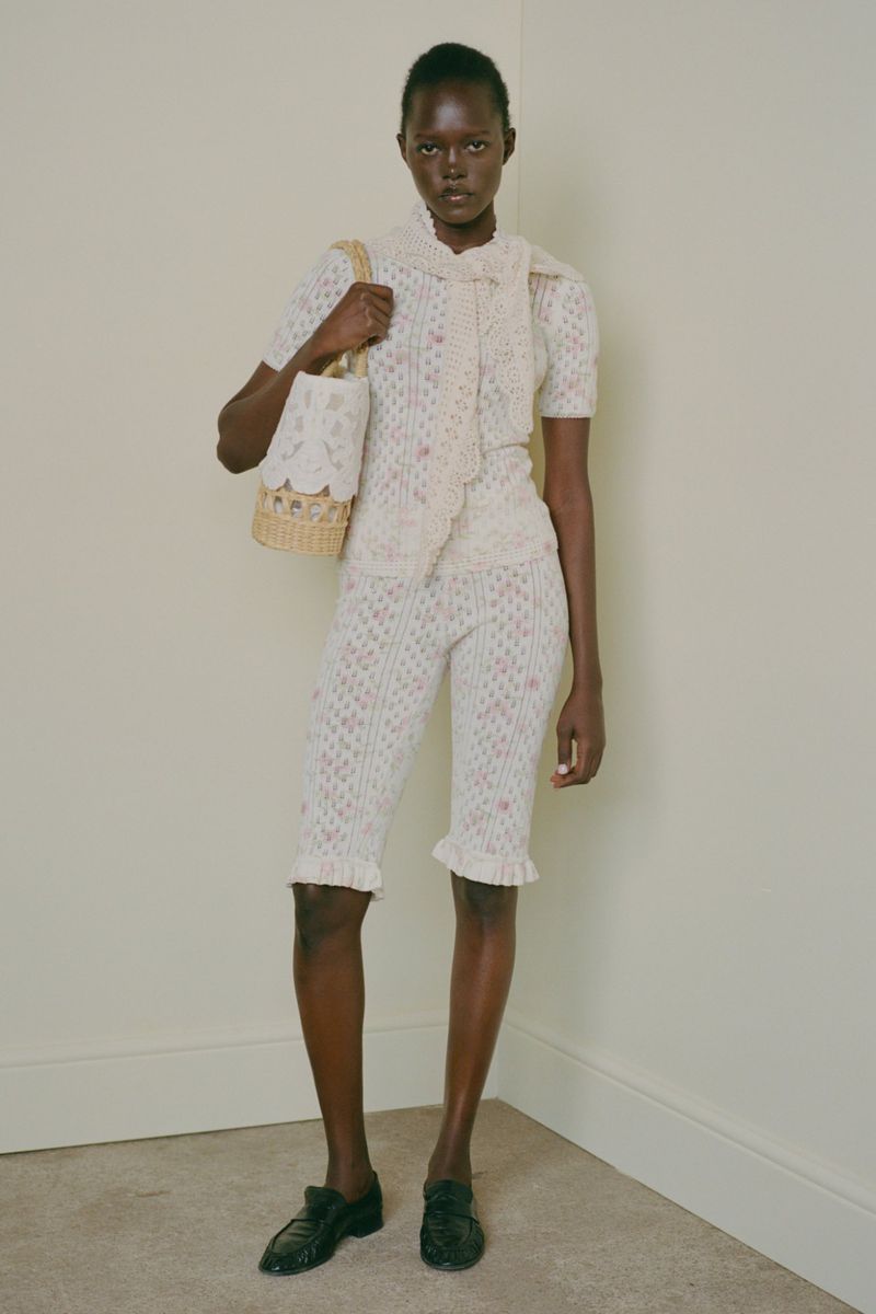 Model wearing patterned knit top, matching shorts, and scarf, holding a woven basket bag against a plain wall.