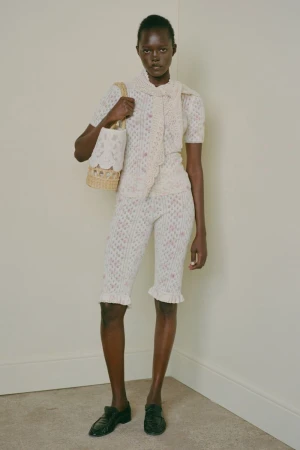 Model wearing patterned knit top, matching shorts, and scarf, holding a woven basket bag against a plain wall.