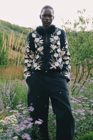 Model wearing a dark embroidered floral jacket and dark trousers standing among tall grass and wildflowers outdoors.