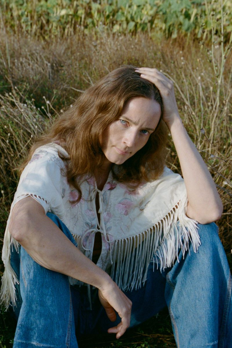Model with long brown hair wearing a fringed embroidered white top and blue jeans seated in a grassy meadow.