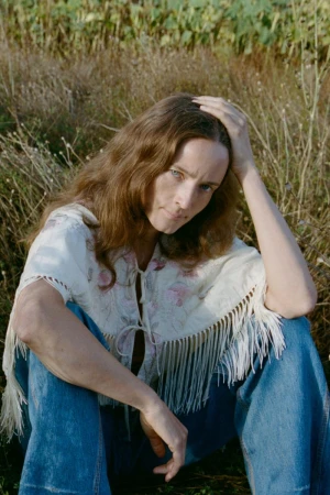 Model with long brown hair wearing a fringed embroidered white top and blue jeans seated in a grassy meadow.