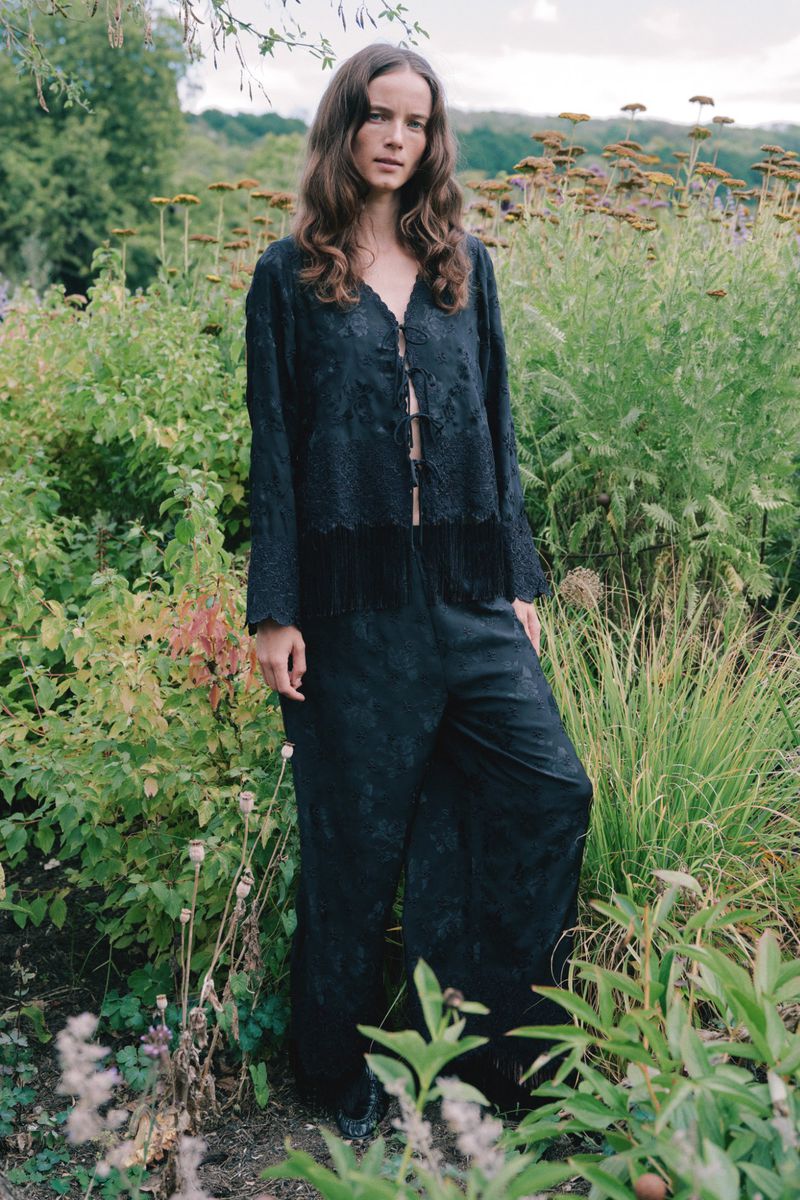 Model standing outdoors amidst tall garden foliage wearing a black patterned long-sleeve top and trousers from Sea SS27 lookbook.