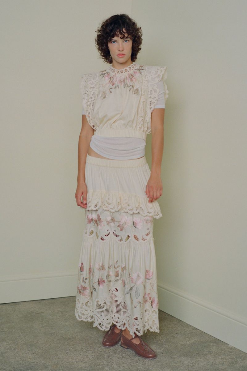 Model with dark curly hair wearing an off-white embroidered top and tiered skirt against a neutral studio wall.