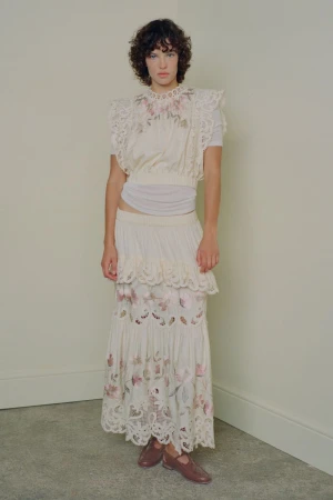 Model with dark curly hair wearing an off-white embroidered top and tiered skirt against a neutral studio wall.