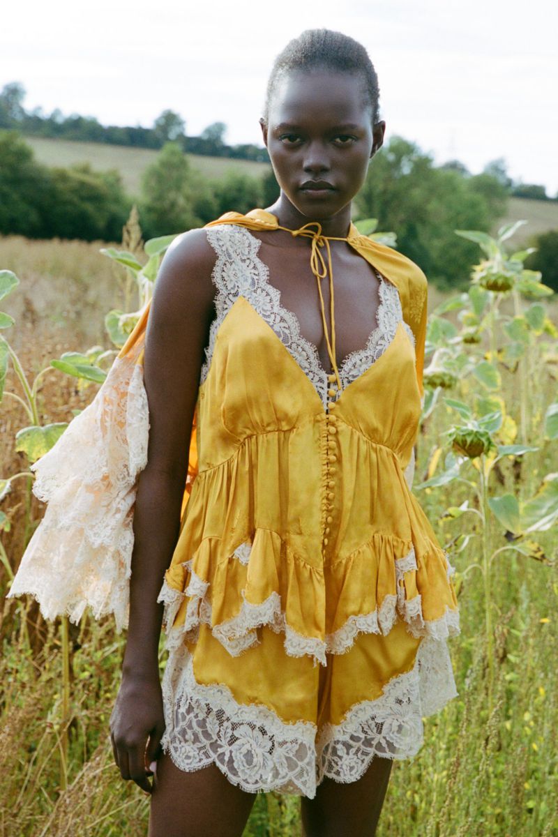 Model wearing a yellow silk slip dress with white lace trim standing outdoors in a tall grassy field.