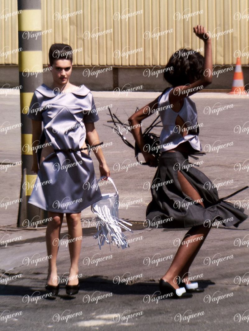 Two models in silver dresses outdoors on pavement with industrial background details, photographed by Dexter Lander.