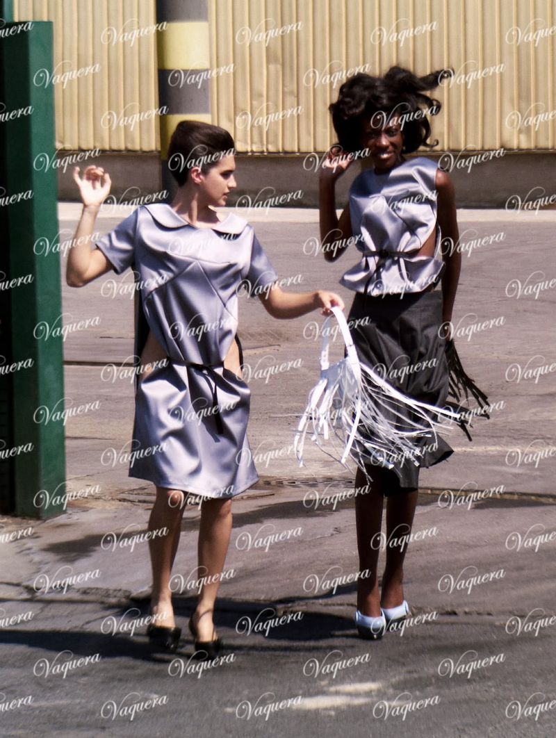 Two models wearing avant-garde metallic grey dresses walking outdoors against an industrial backdrop for Vaquera FW26.