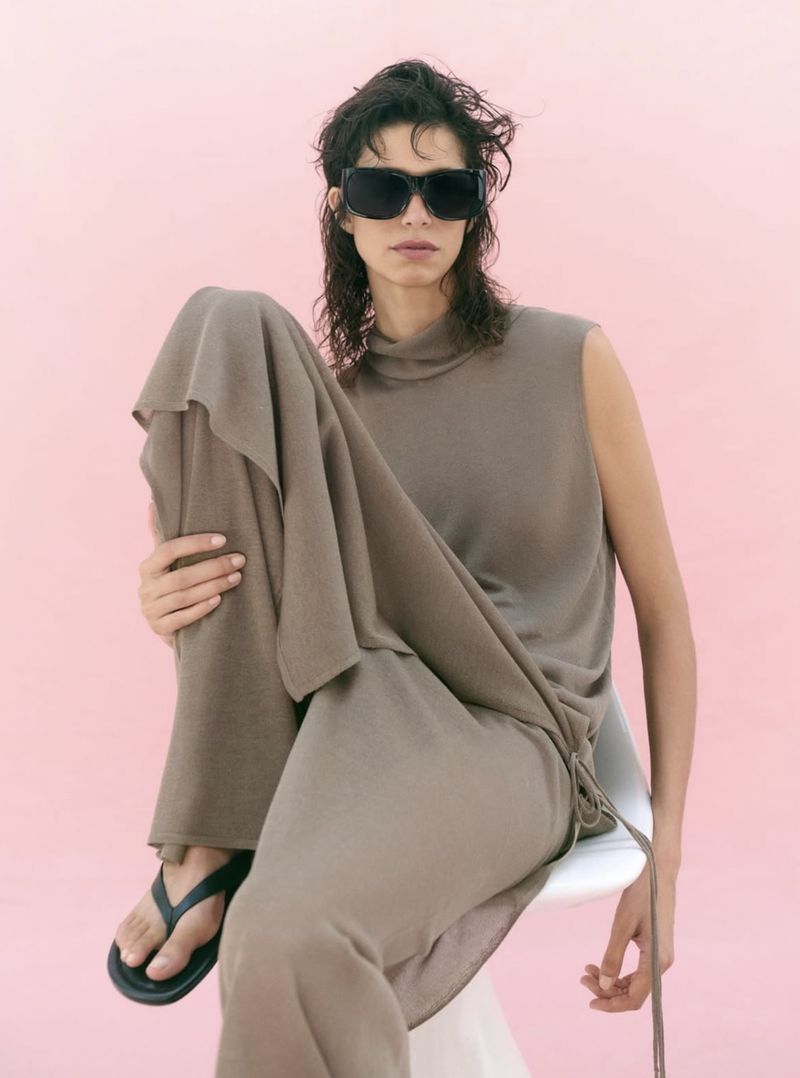 Model Mica Argañaraz wearing a brown knit dress and sunglasses seated on a white chair against a pink backdrop for Zara.