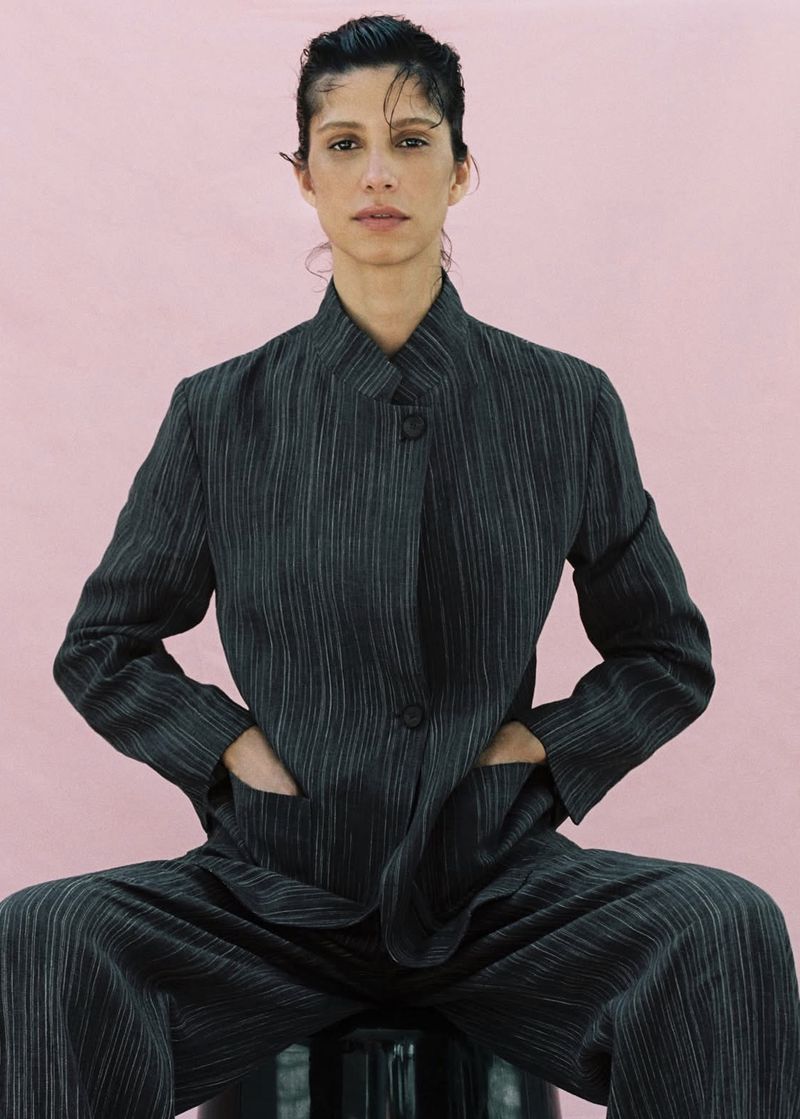 Model Mica Argañaraz wearing a dark pinstripe suit jacket against a pink backdrop