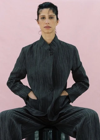 Model Mica Argañaraz wearing a dark pinstripe suit jacket against a pink backdrop