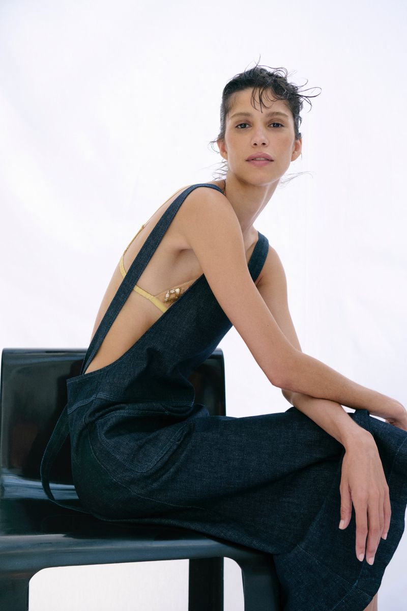 Model Mica Argañaraz wearing dark denim overalls seated on a dark chair against a white background for Zara.