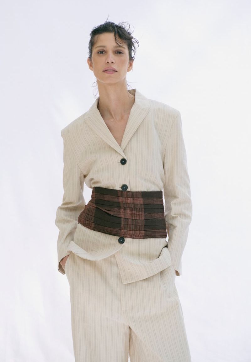 Model Mica Argañaraz wearing a cream blazer and trousers with a wide patterned cummerbund against white.